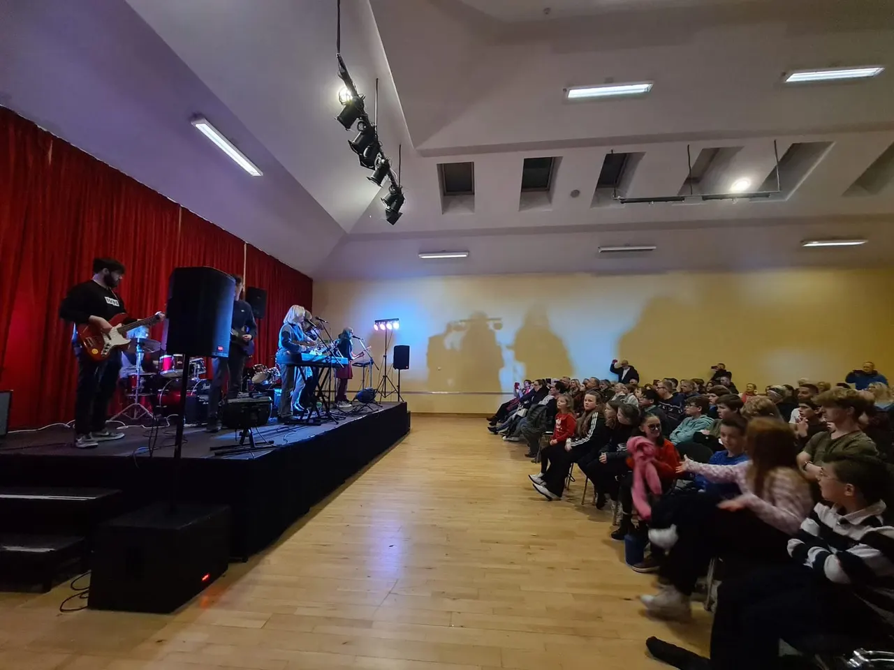 A live band performs on an indoor stage with red curtains as an audience of children sits on the floor watching the performance.