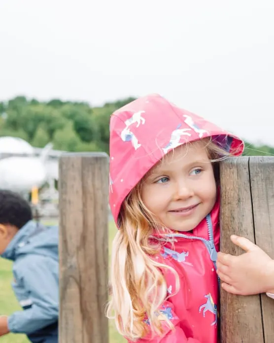 Young child in pink patterned raincoat peering over wooden fence at rural venue.