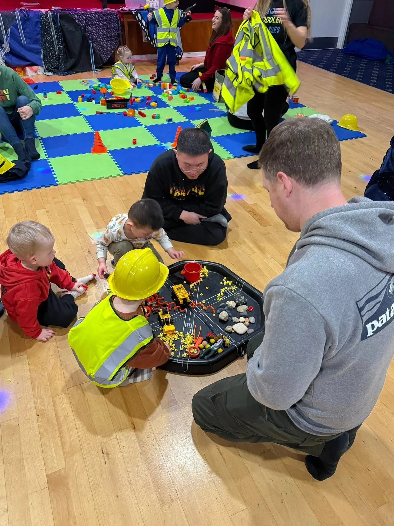 Young children and adults participate in a construction-themed activity session indoors, wearing hard hats and high-visibility vests.