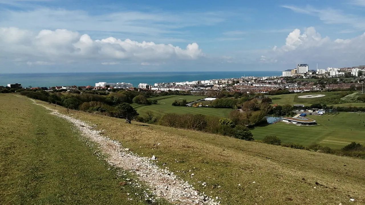 Green downland with chalky path sloping toward a coastal town, sea and leisure facilities nestled in valley below.