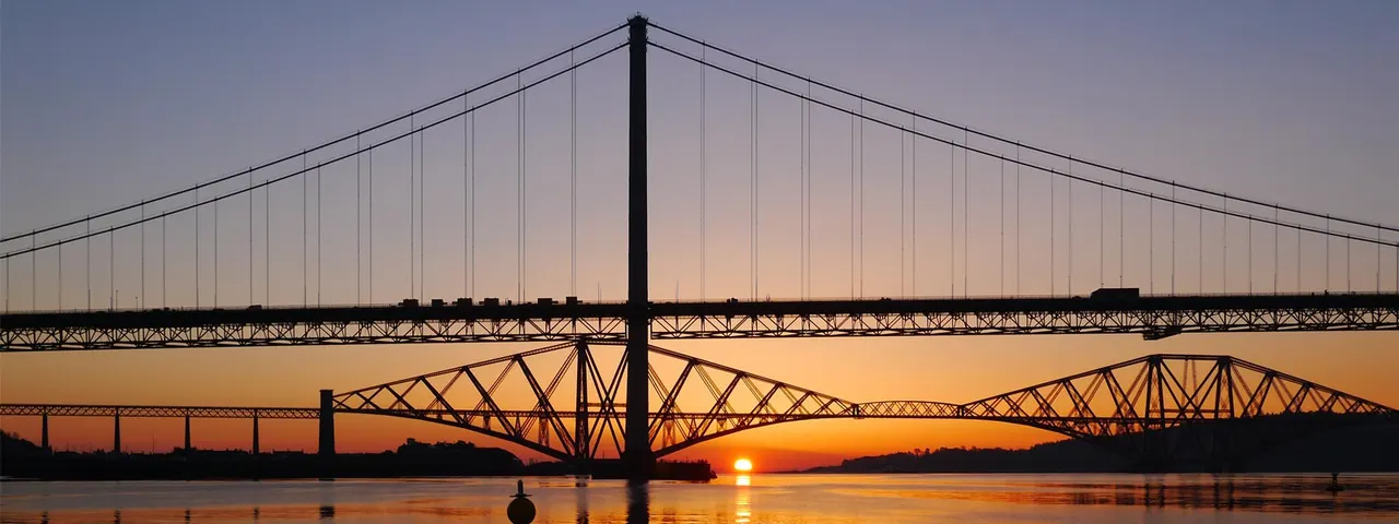Two suspension bridges silhouetted against an orange sunset over water.