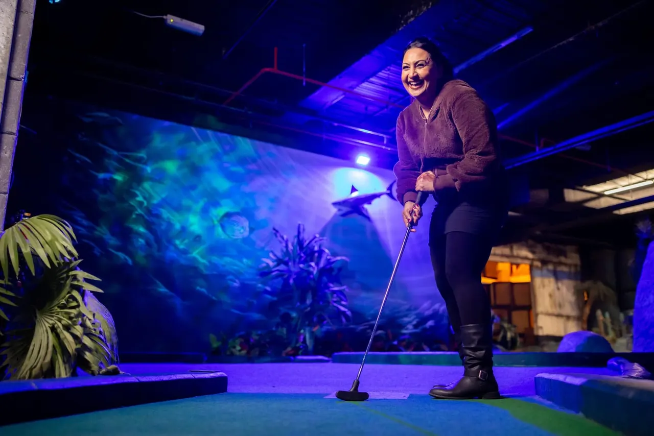 Woman playing minigolf indoors under blue lighting with themed aquatic backdrop and decorative plants.