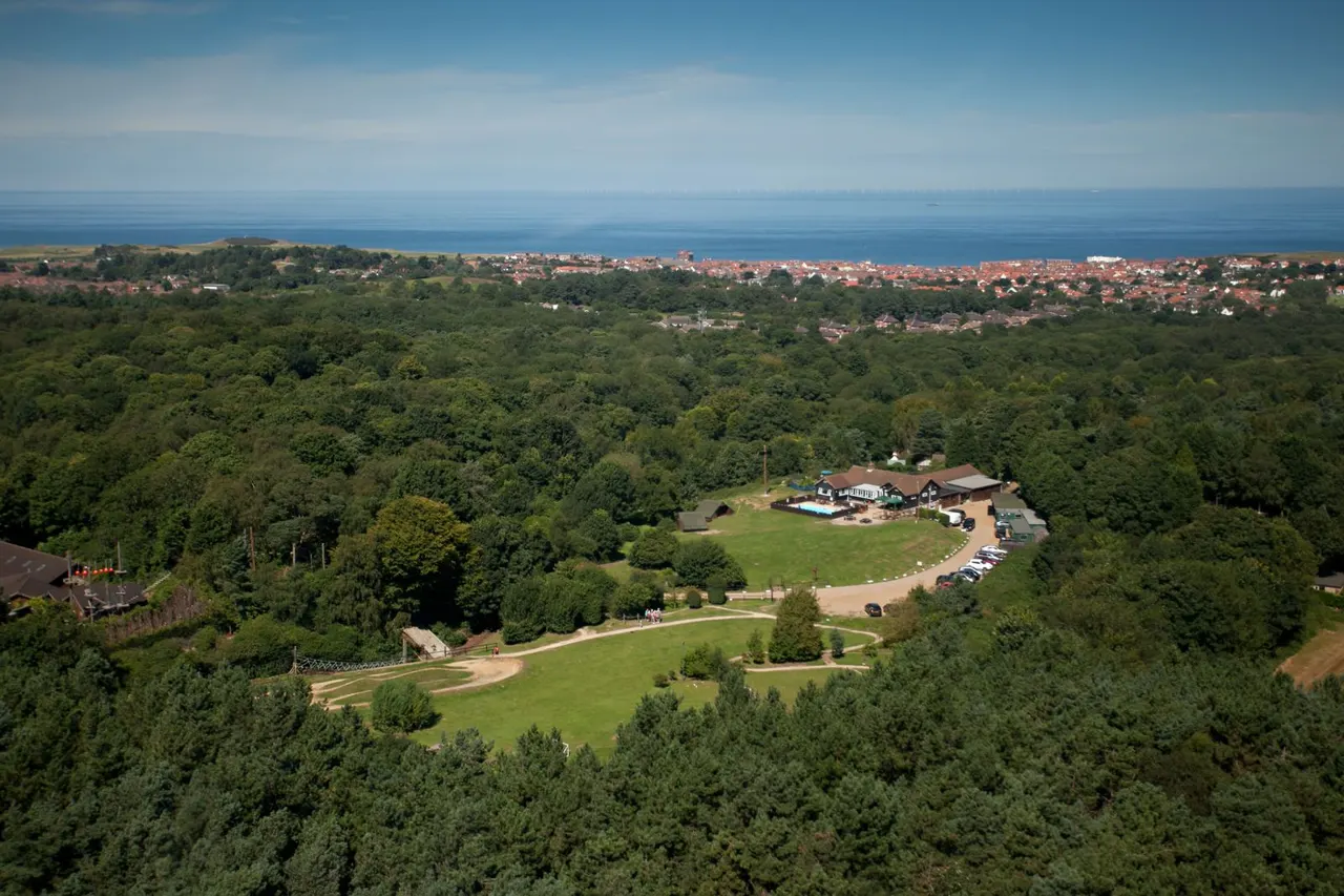 Aerial view of a leisure centre with pool and grounds surrounded by woodland, overlooking coastal town and sea.