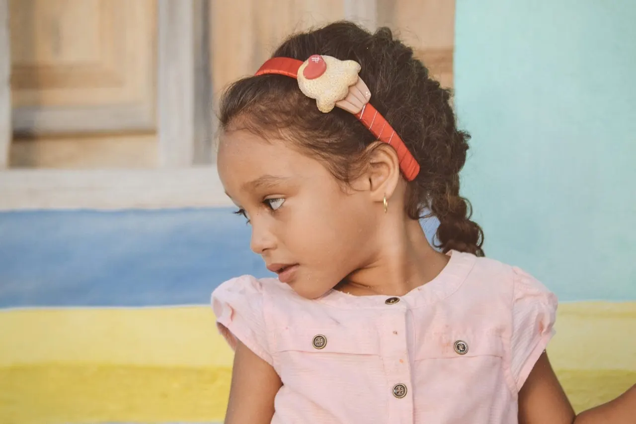 Young child wearing a red hair accessory and pink top, seated against a colourful striped background.