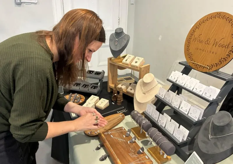 Woman examining jewellery displayed on wooden stands and cards at a craft market stall.