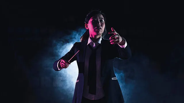 A conductor in formal attire gestures expressively whilst holding a baton against dramatic blue and pink stage lighting.