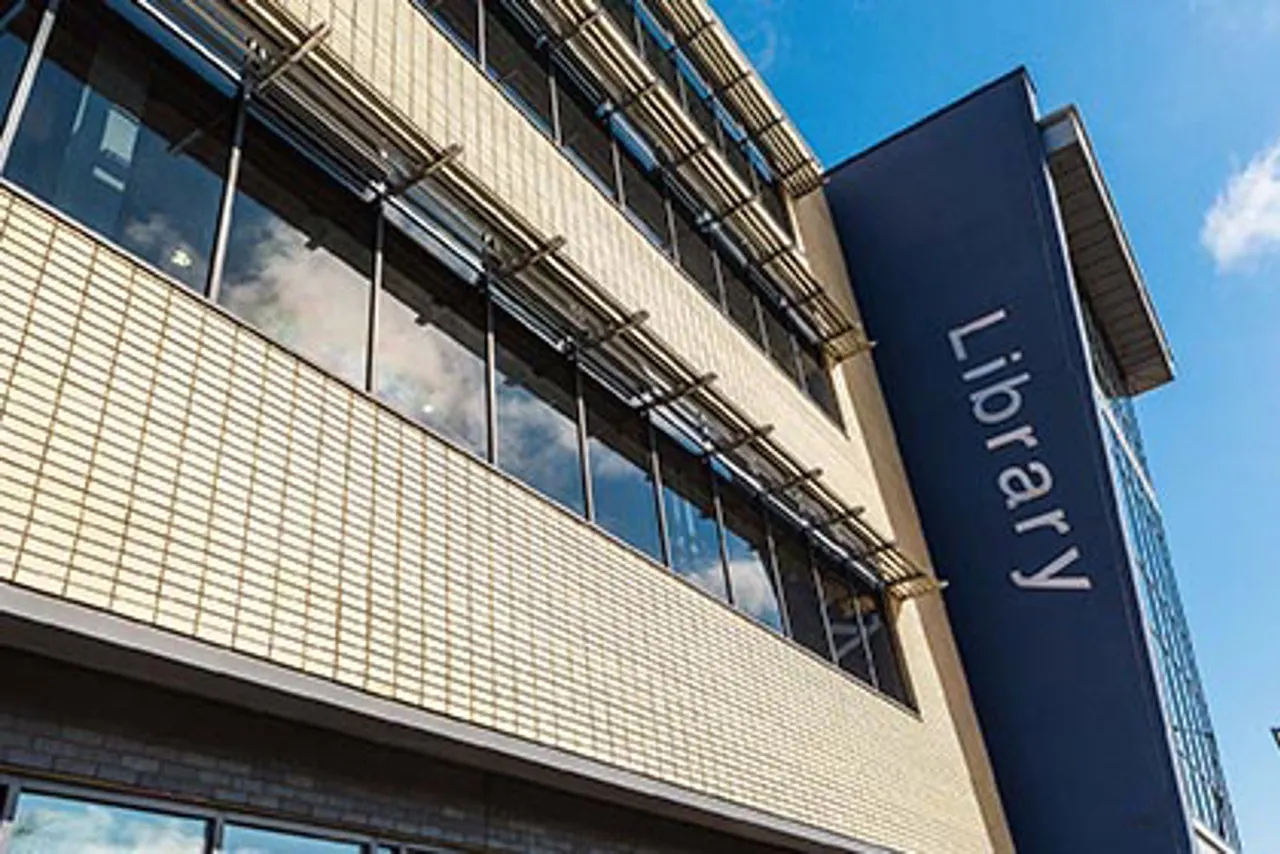 Exterior of modern library building with cream brick façade, large windows, and blue vertical signage against clear sky.