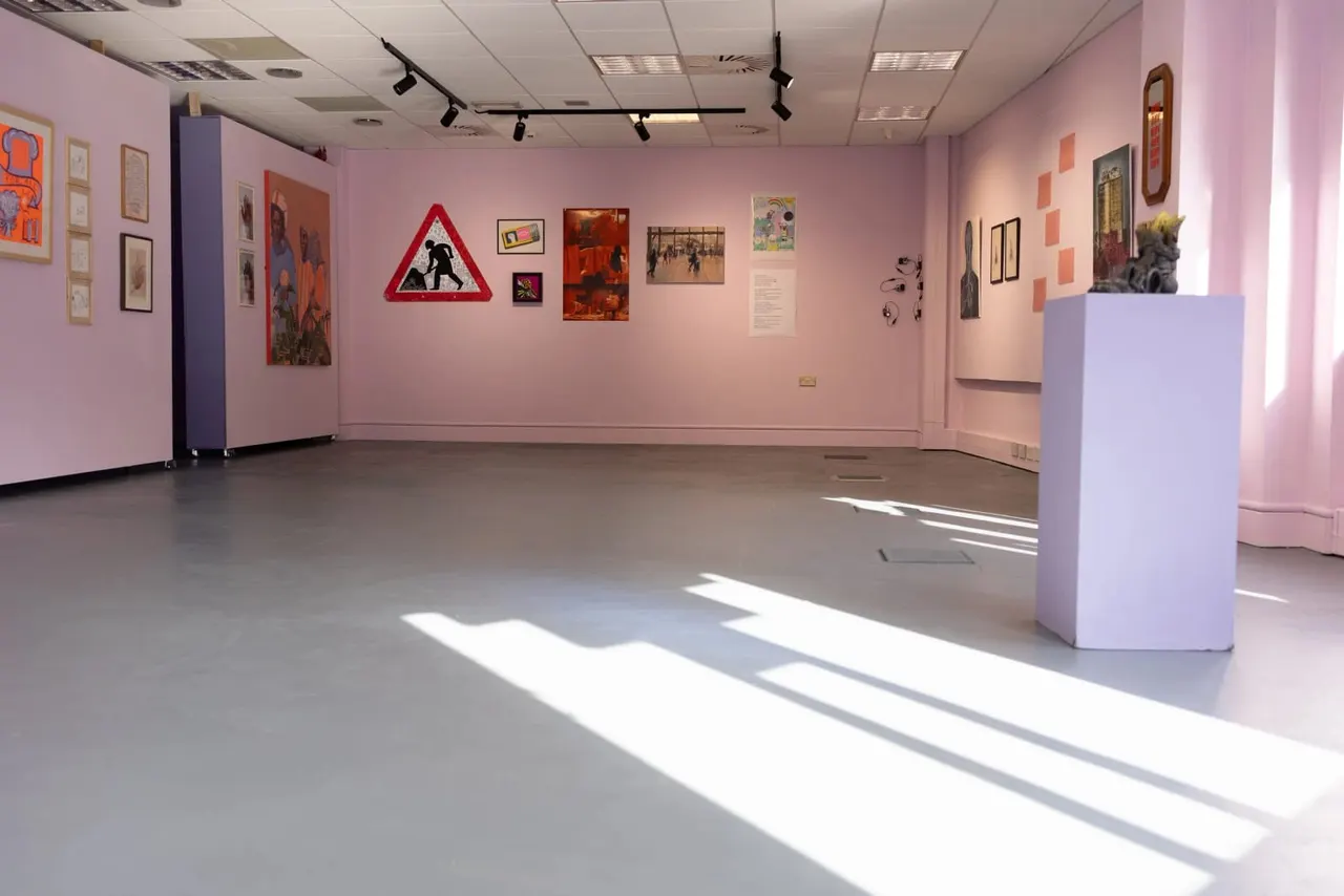 Interior of an art gallery with pink walls displaying various artworks and a red triangular warning sign, with a white plinth in the centre and sunlight streaming across the floor.