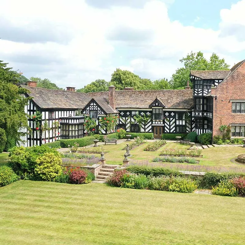 Tudor and brick manor house with formal gardens and manicured lawns under cloudy sky.