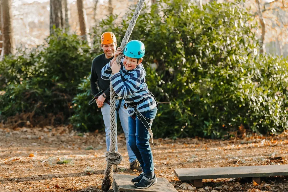 A child in a blue helmet climbs a rope obstacle while an adult supervises during an outdoor activity course.