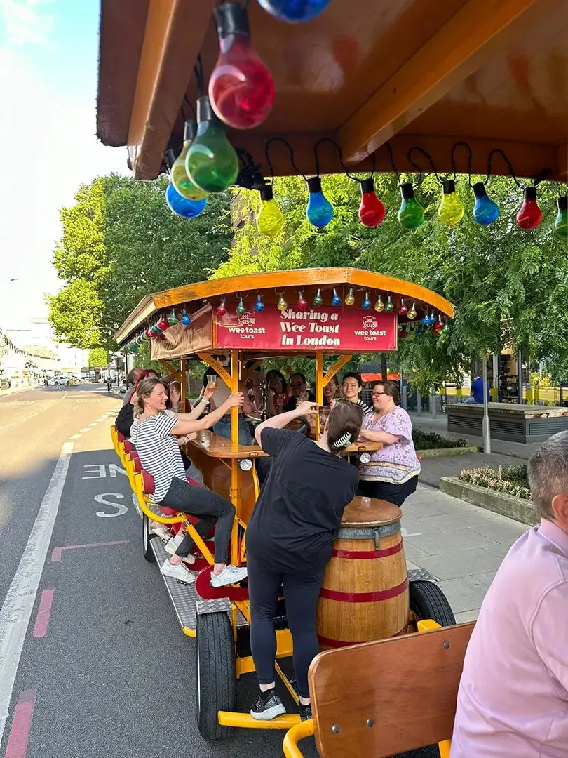 A group of people pedalling a colourful pub bike along a London street, decorated with hanging coloured lights.
