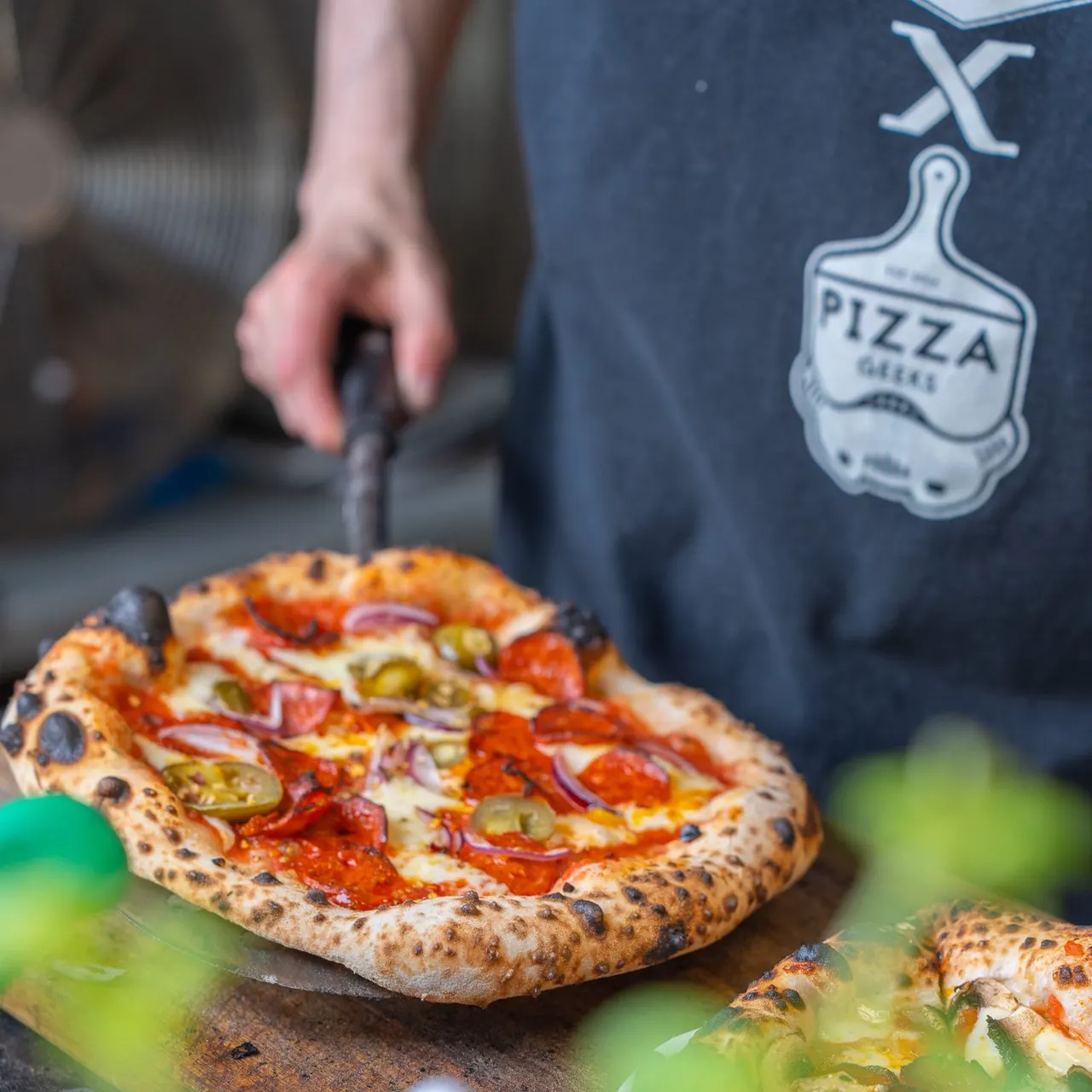 A person in a pizza restaurant uniform holds a pizza cutter over a freshly made wood-fired pizza topped with pepperoni and peppers.