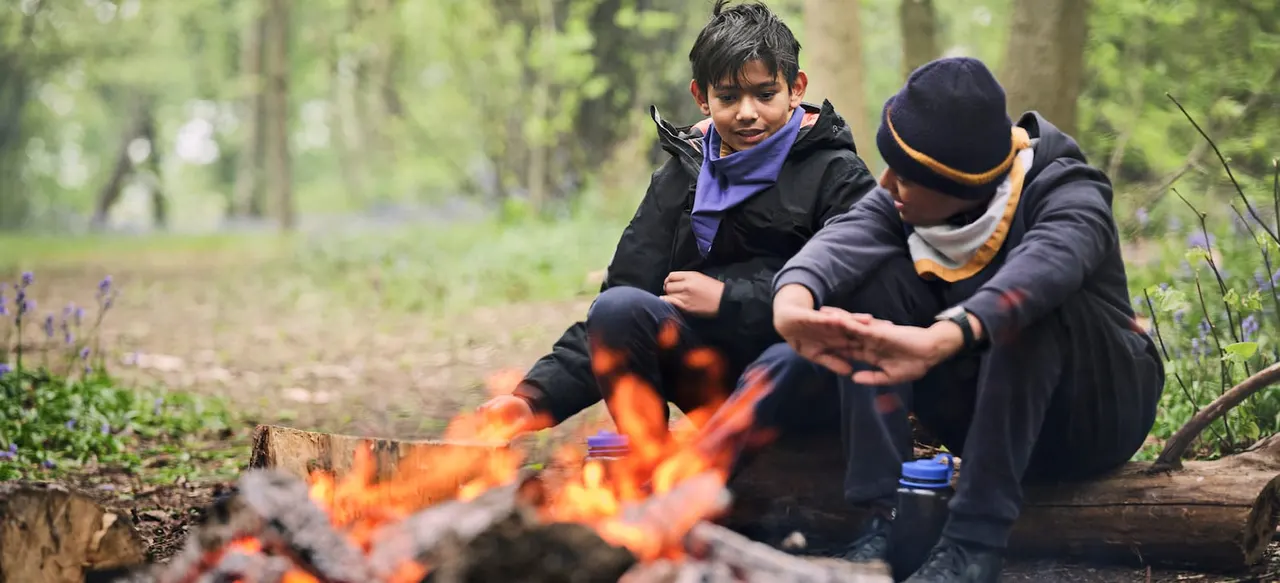Two young scouts warming themselves by a campfire in a wooded clearing.