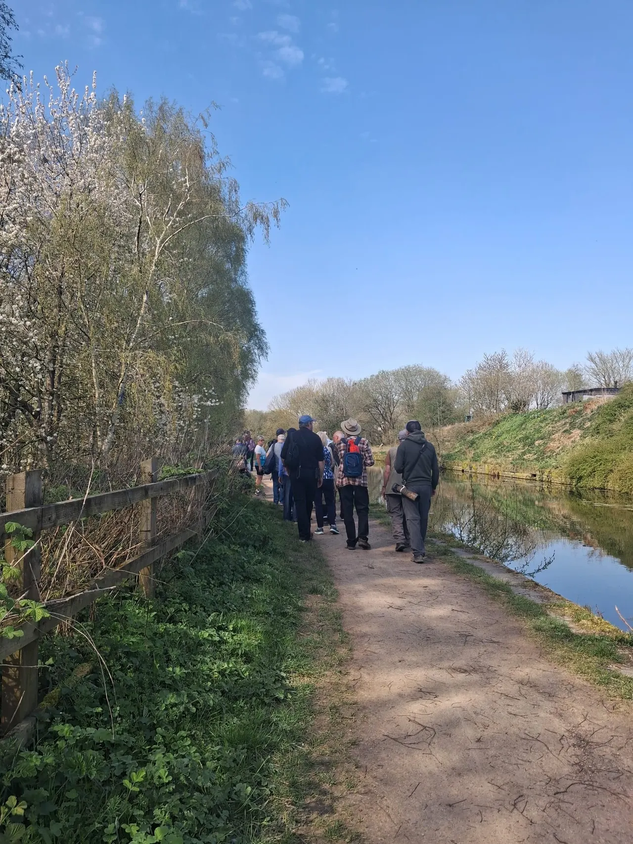 A group of visitors walking along a towpath beside a canal lined with budding trees on a clear spring day.