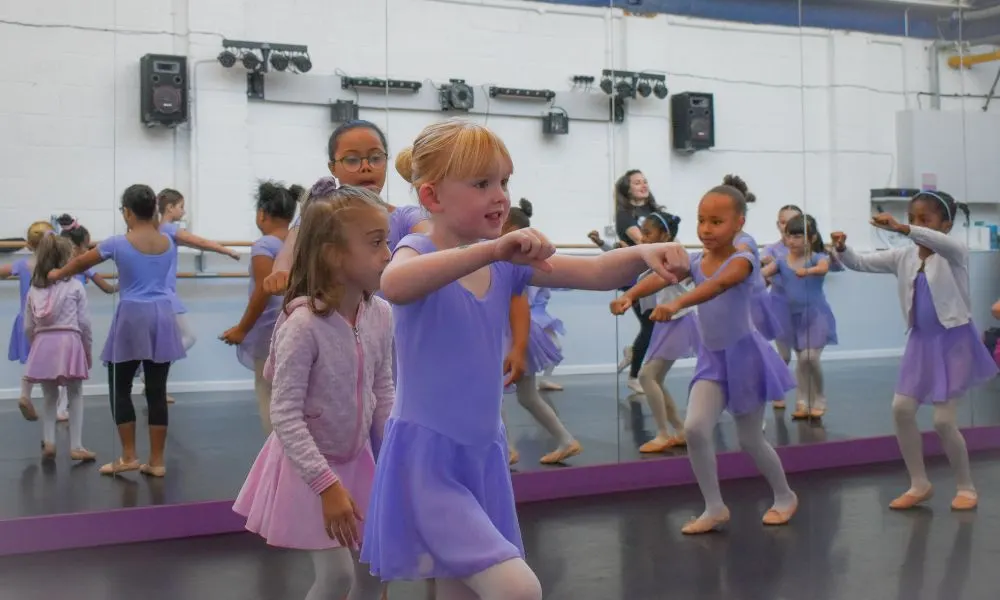 Group of young children in purple and pink dancewear performing choreography in a mirrored studio.