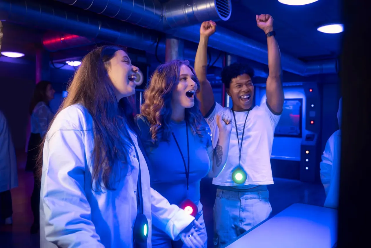 Three people wearing illuminated armbands celebrate and cheer in a blue-lit indoor gaming arena.