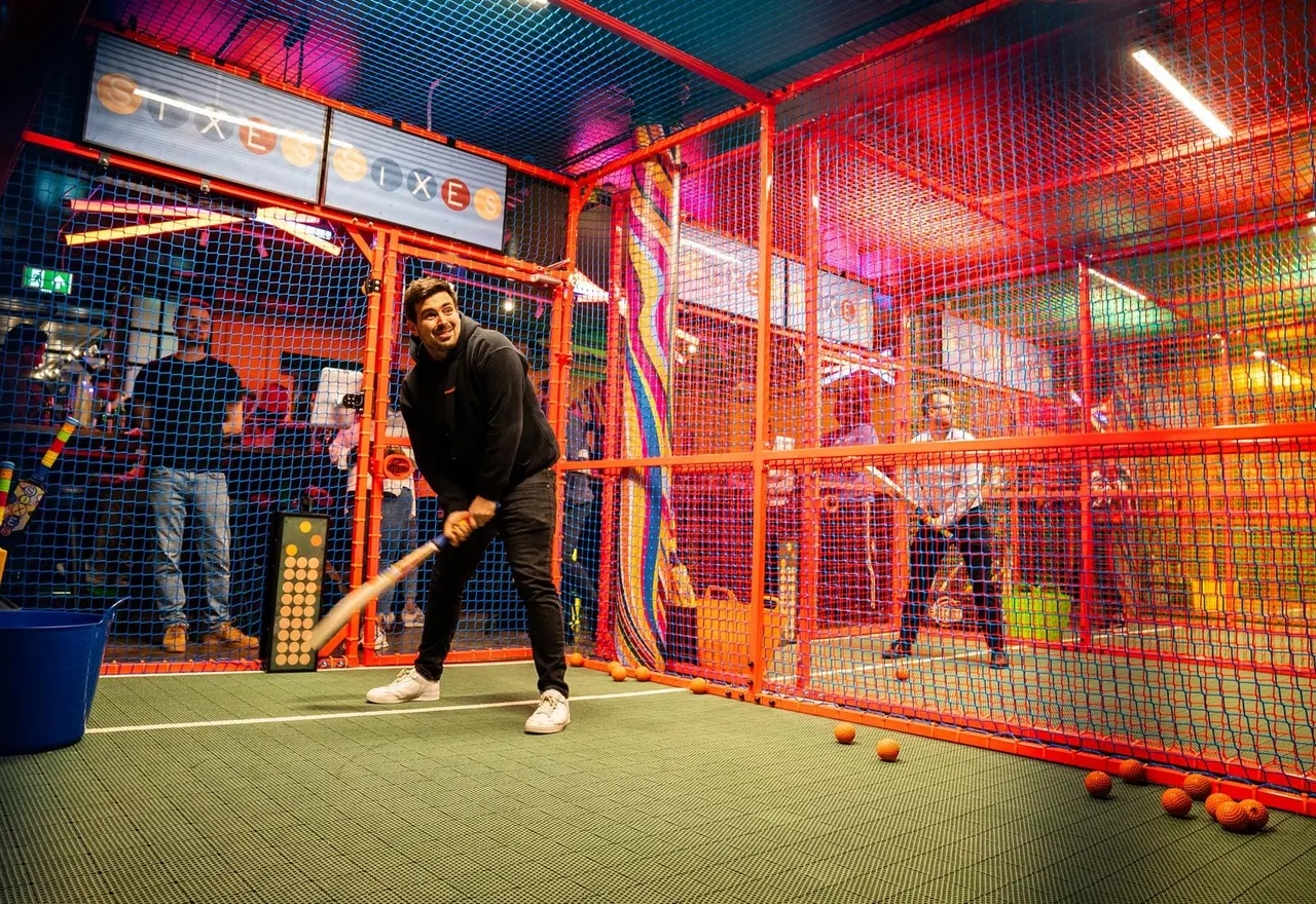 Man batting in an indoor cricket cage with red netting and neon lighting.