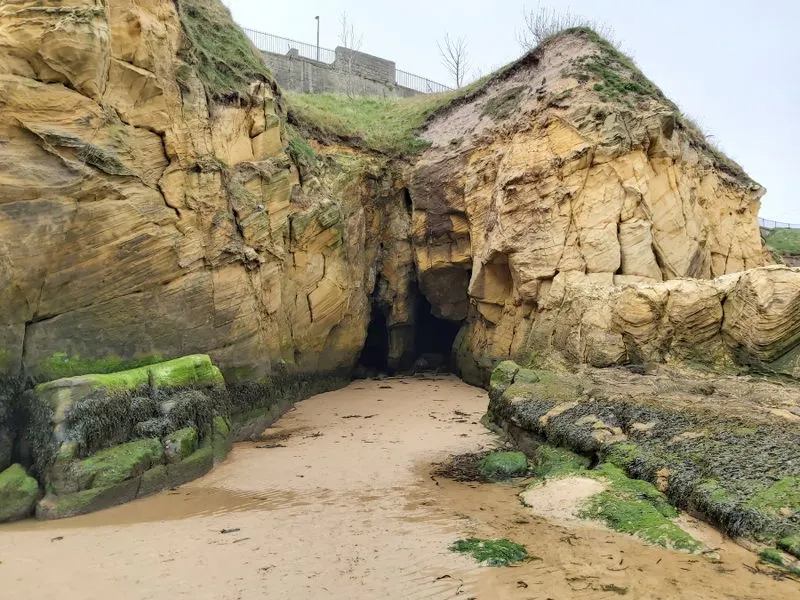 Sandy beach cove with large natural rock cave entrance flanked by moss-covered stone formations and cliffs.