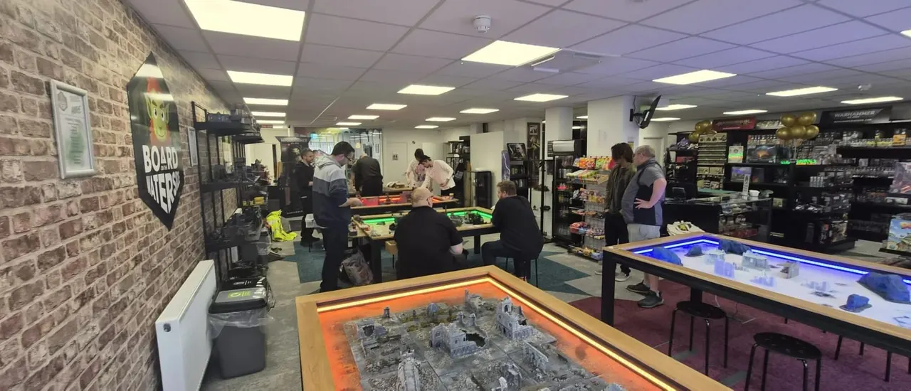 Interior of a board game café with visitors playing games at tables, display cases, and gaming boards visible throughout the space.