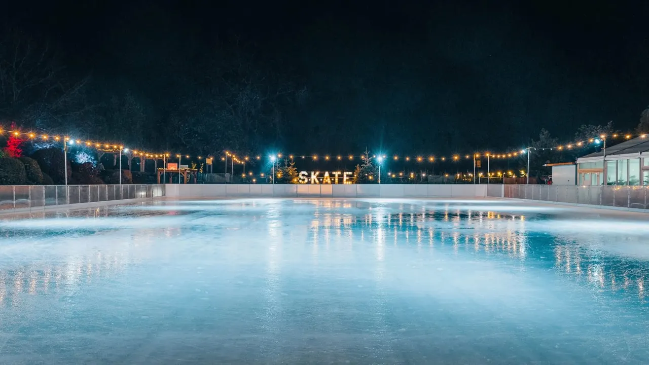 Empty outdoor ice skating rink at night with illuminated "SKATE" sign and fairy lights strung overhead.