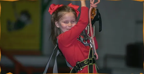 Child wearing red jacket and red hair bows on an indoor climbing wall with safety harness.