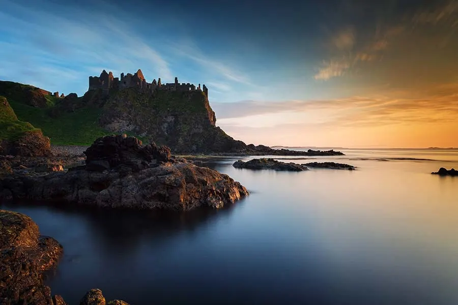 Ruined castle silhouetted on rocky headland at sunset, with calm sea and rocky coastline in foreground.