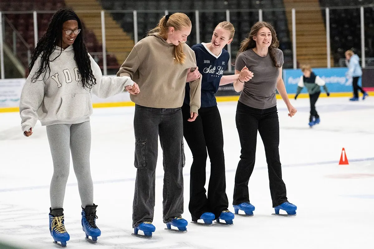 Four young women ice skating together in a line, holding hands and smiling on an indoor ice rink.