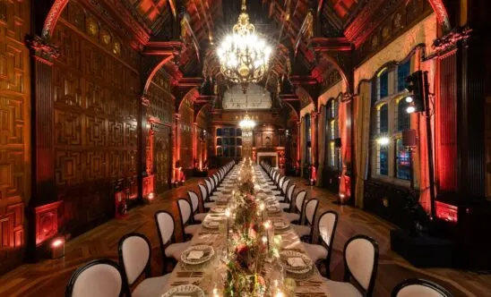 Long formal dining table set for a banquet in an ornate red and gold historic hall with elaborate chandelier.