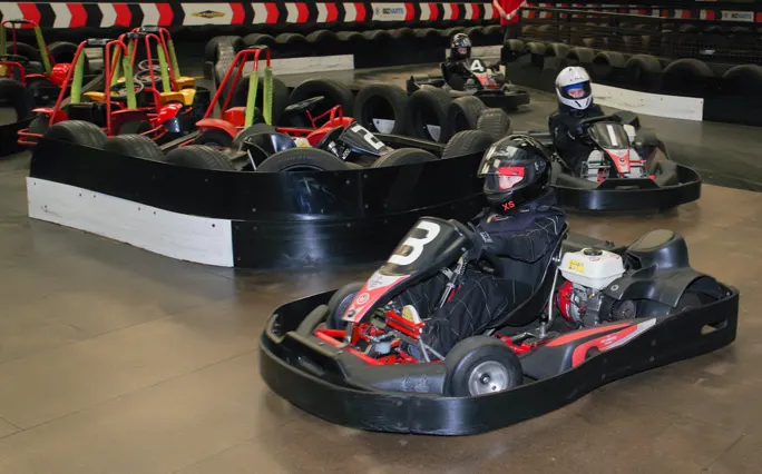 Multiple drivers in helmets racing go-karts on an indoor track with tyre barriers and striped safety barriers visible.