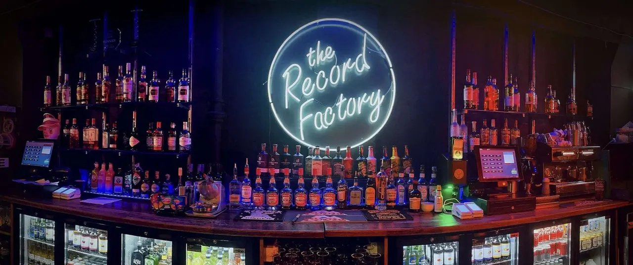 Bar counter lined with spirits bottles and illuminated neon "The Record Factory" sign above.