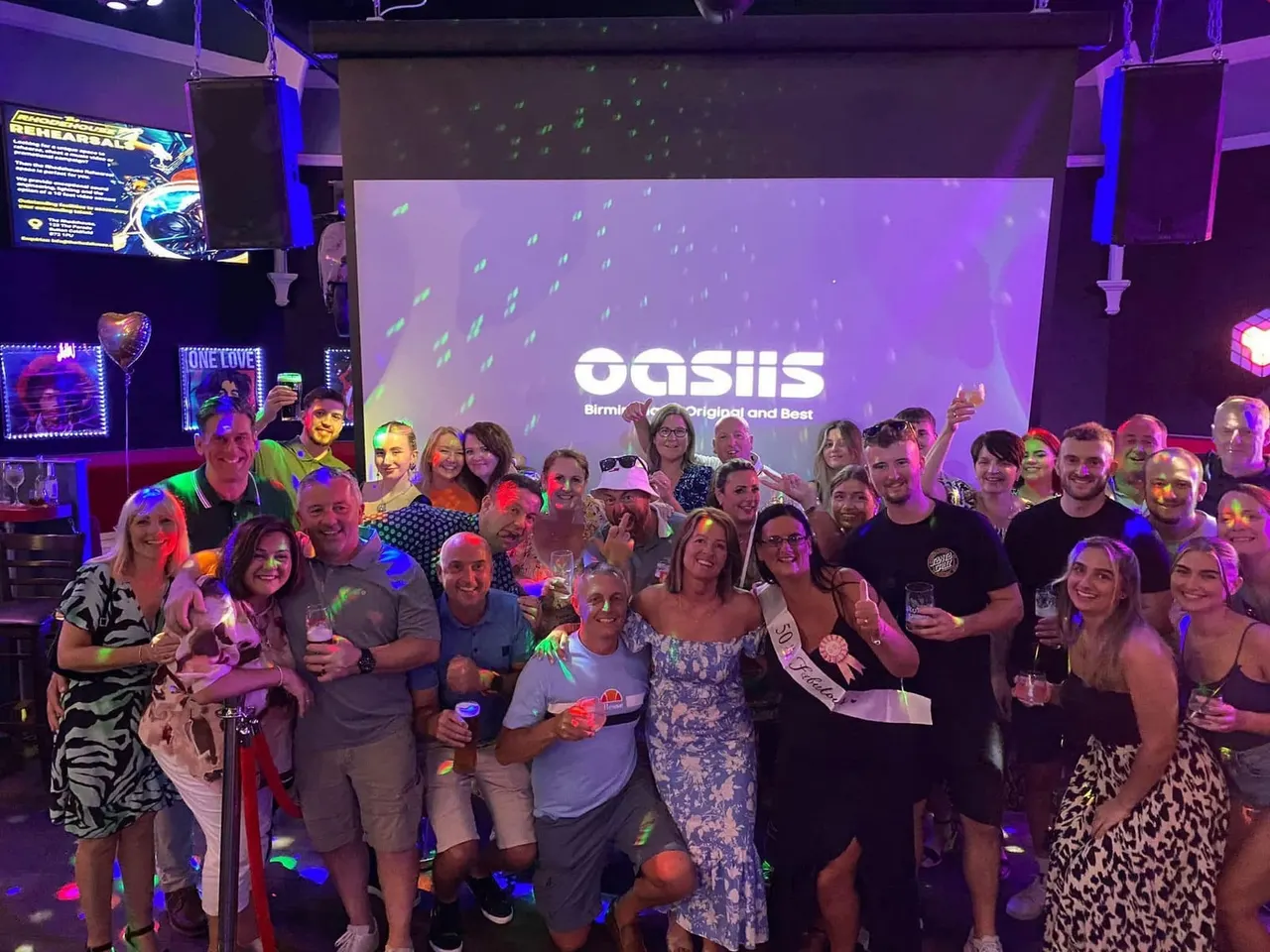 Large group of people posing together in a nightclub with purple lighting, holding drinks and smiling at the camera.