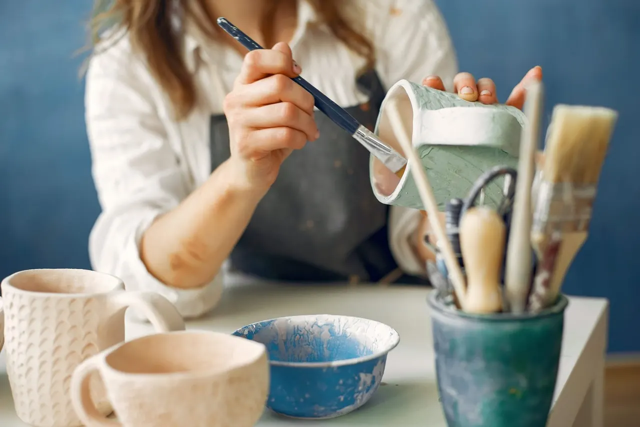 Person in apron holding paintbrush and fabric during pottery or craft painting activity at a table.