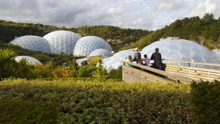 Visitors on a wooden platform overlook large white geodesic domes nestled in a verdant valley surrounded by woodland.