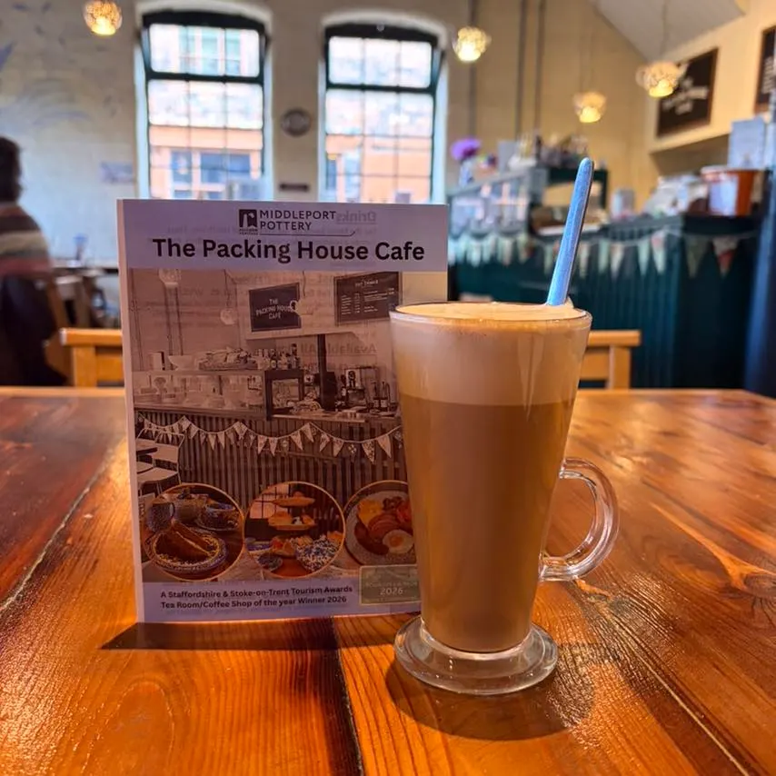 A latte with latte art sits next to a menu for The Packing House Cafe on a wooden table inside a bright, airy café.