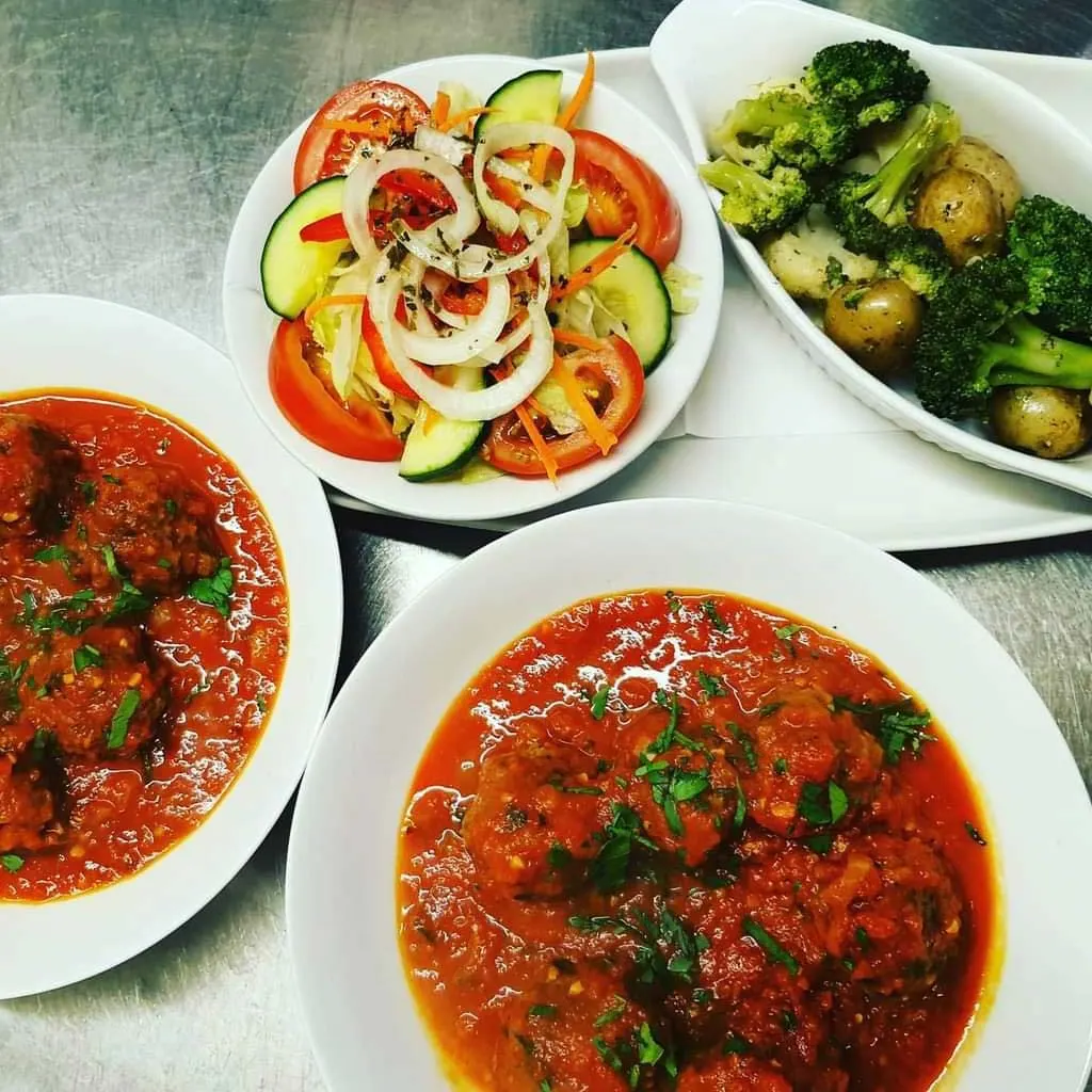 Multiple white plates of Italian food including red stews, fresh vegetable salad, and roasted broccoli and potatoes.