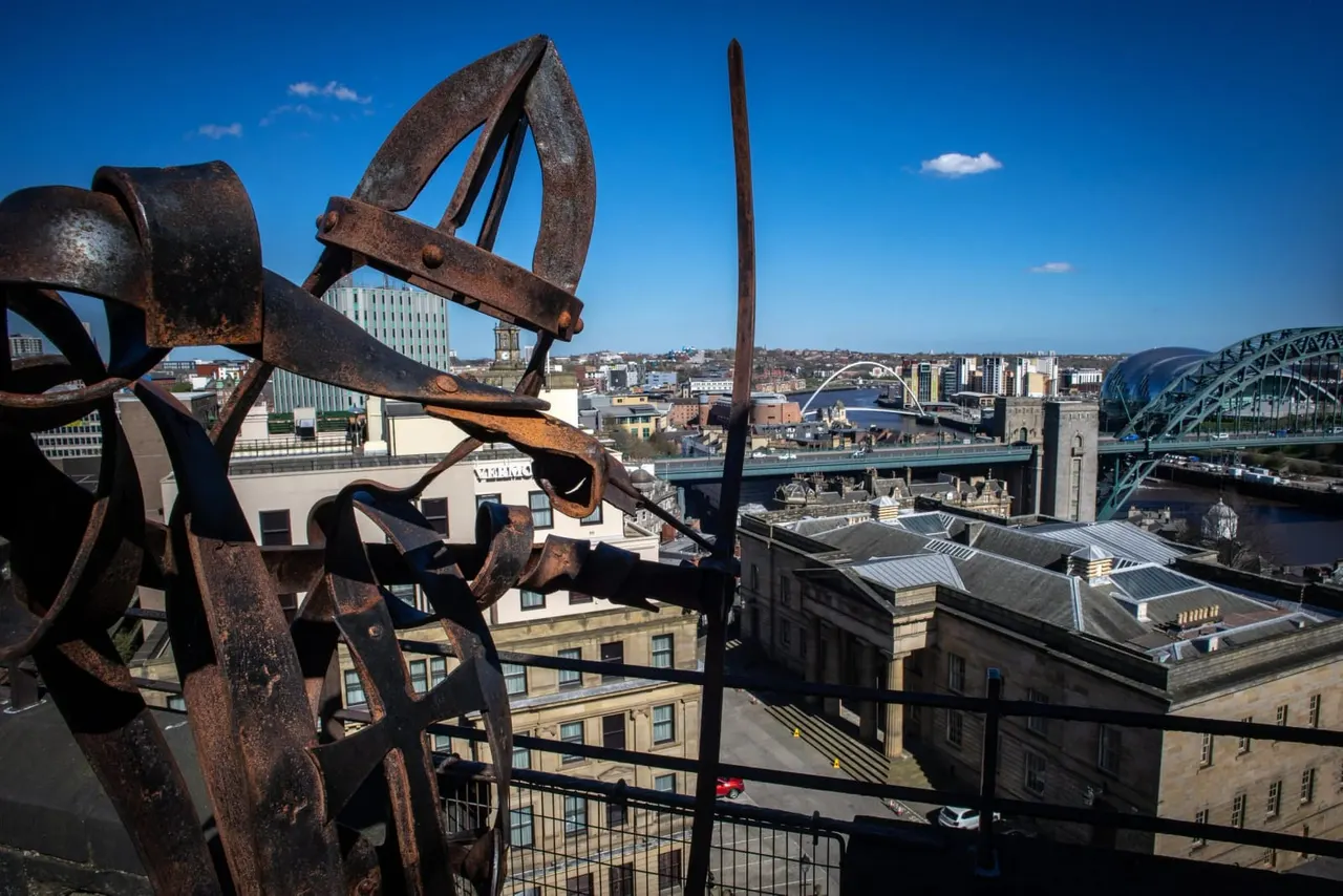 Rusted industrial sculpture in foreground with Newcastle quayside, River Tyne and Tyne Bridge visible beyond under clear blue sky.