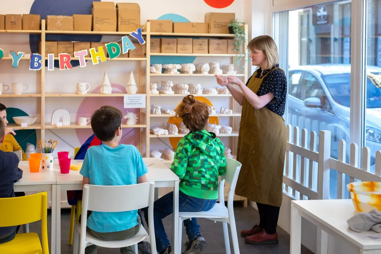 An adult instructor leads a birthday workshop for children seated at a table in a bright pottery studio with shelves of ceramic pieces and a birthday banner.