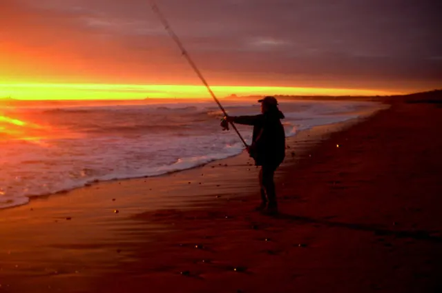 Person fishing from the beach at sunset with golden and orange sky reflected on the water.