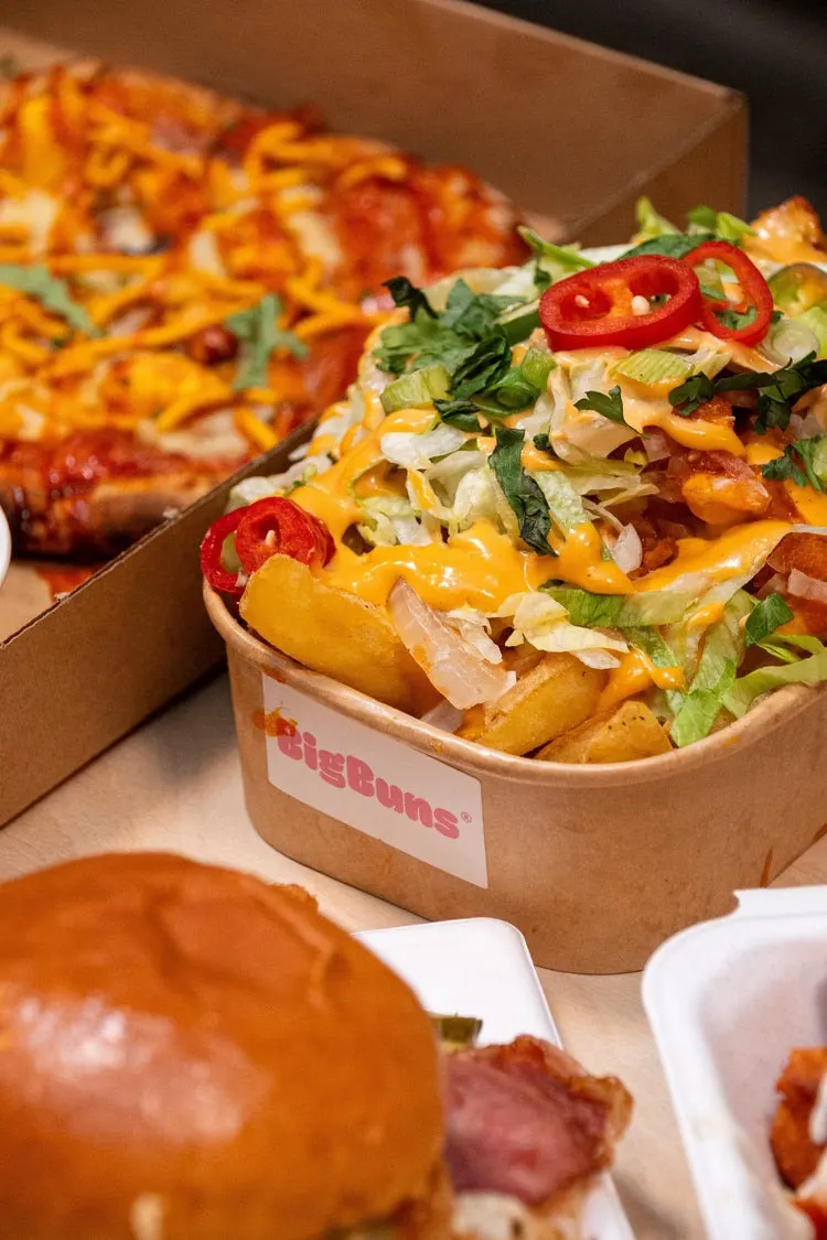 Loaded chips with melted cheese, fresh herbs, red chillies and bacon in a cardboard container, with pizza and burgers nearby.