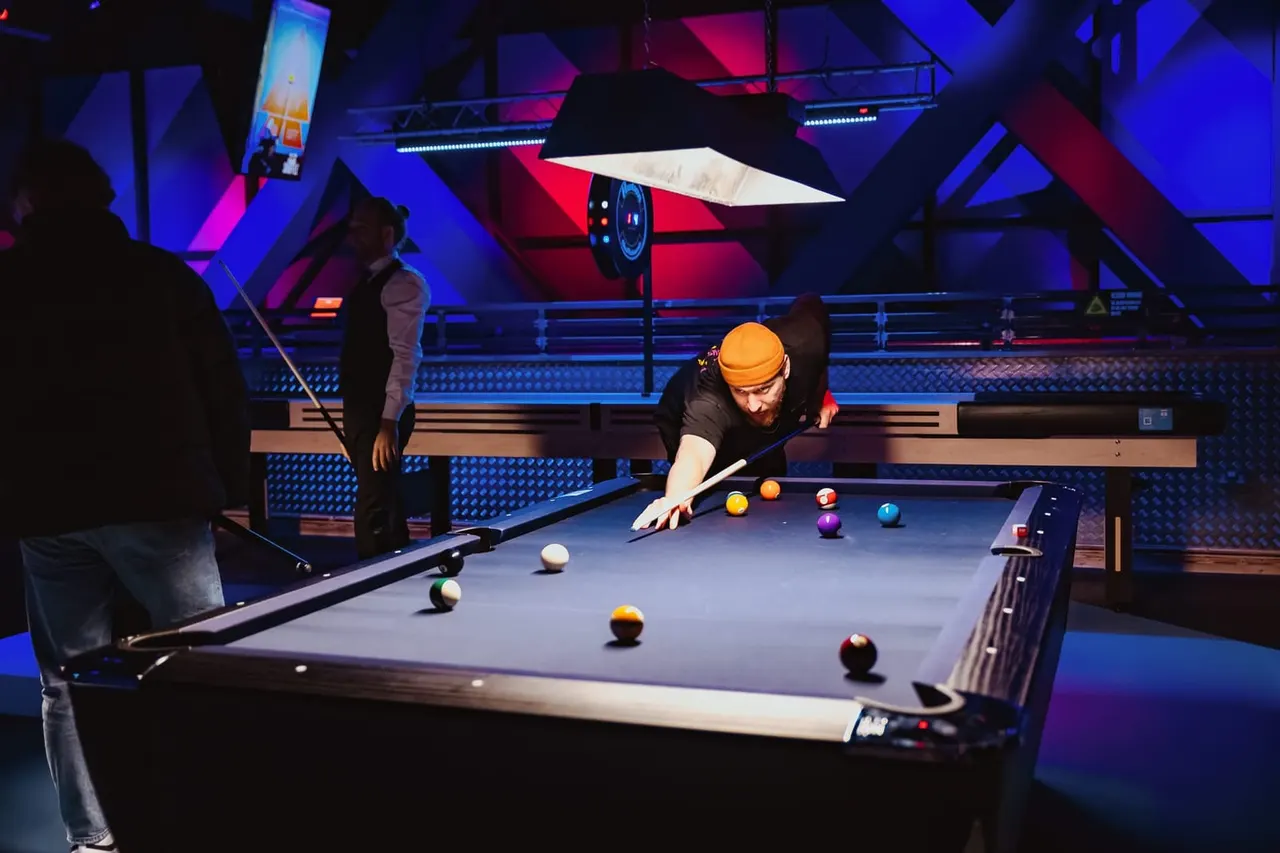Player in yellow beanie leans over pool table taking a shot in a neon-lit arcade venue with spectators nearby.