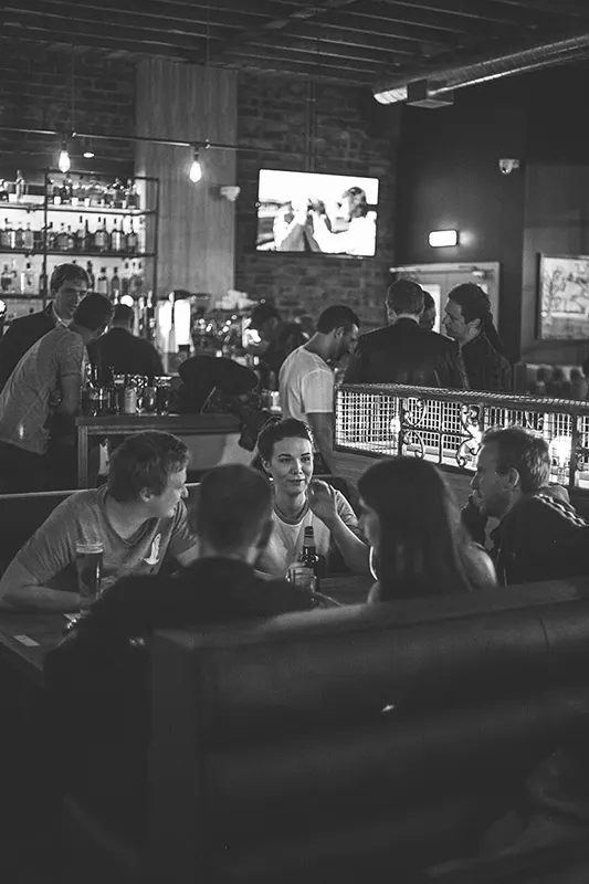 Customers sit at tables in a busy bar with a TV screen and lit bottles displayed behind the counter.