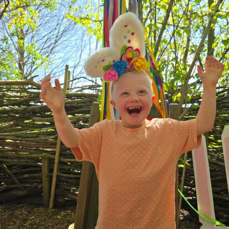 Young child wearing a colourful bunny headdress with arms raised, laughing outdoors among trees.