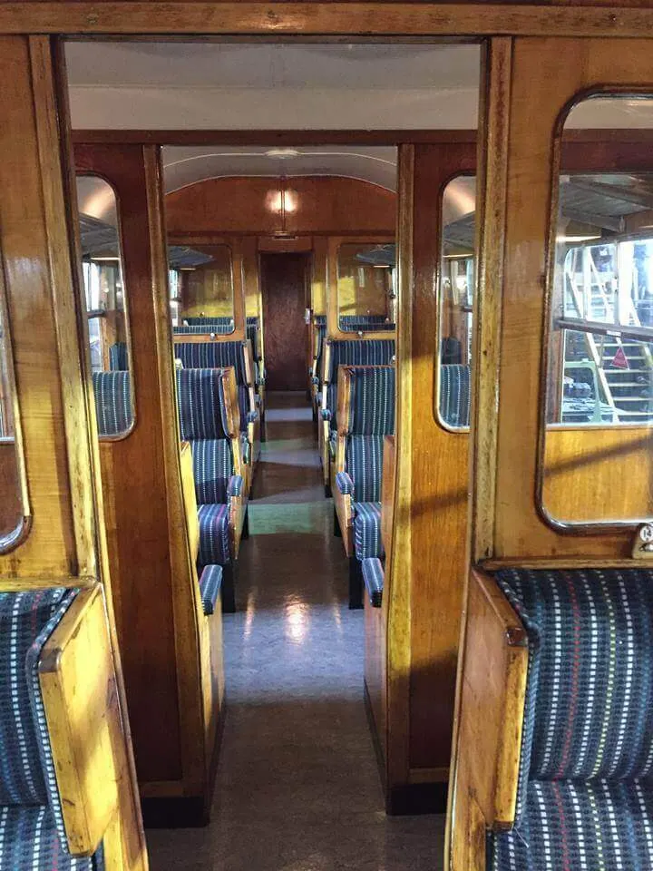 Interior of a vintage railway carriage with wooden panelling, blue striped seats, and natural light from windows along the aisle.