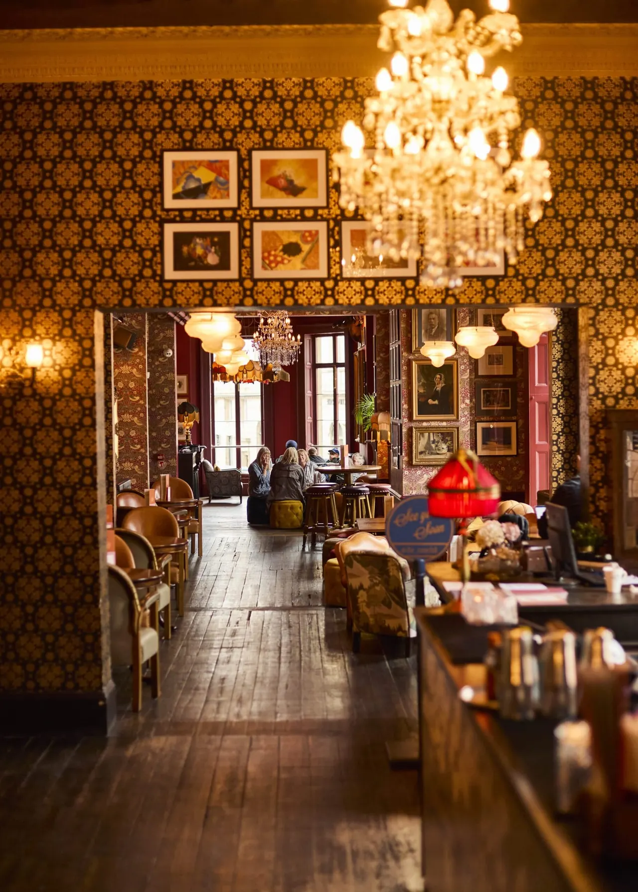 Elegant bar and lounge interior with patterned wallpaper, framed artwork, crystal chandeliers, and guests seated at tables with warm ambient lighting.