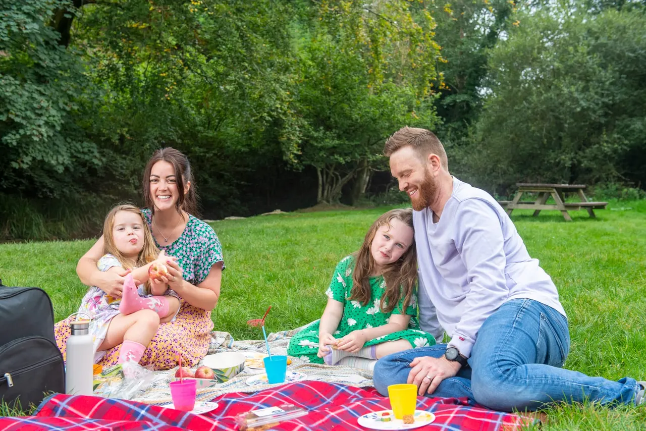 Family of four enjoying a picnic on a red tartan blanket in a grassy park surrounded by trees.