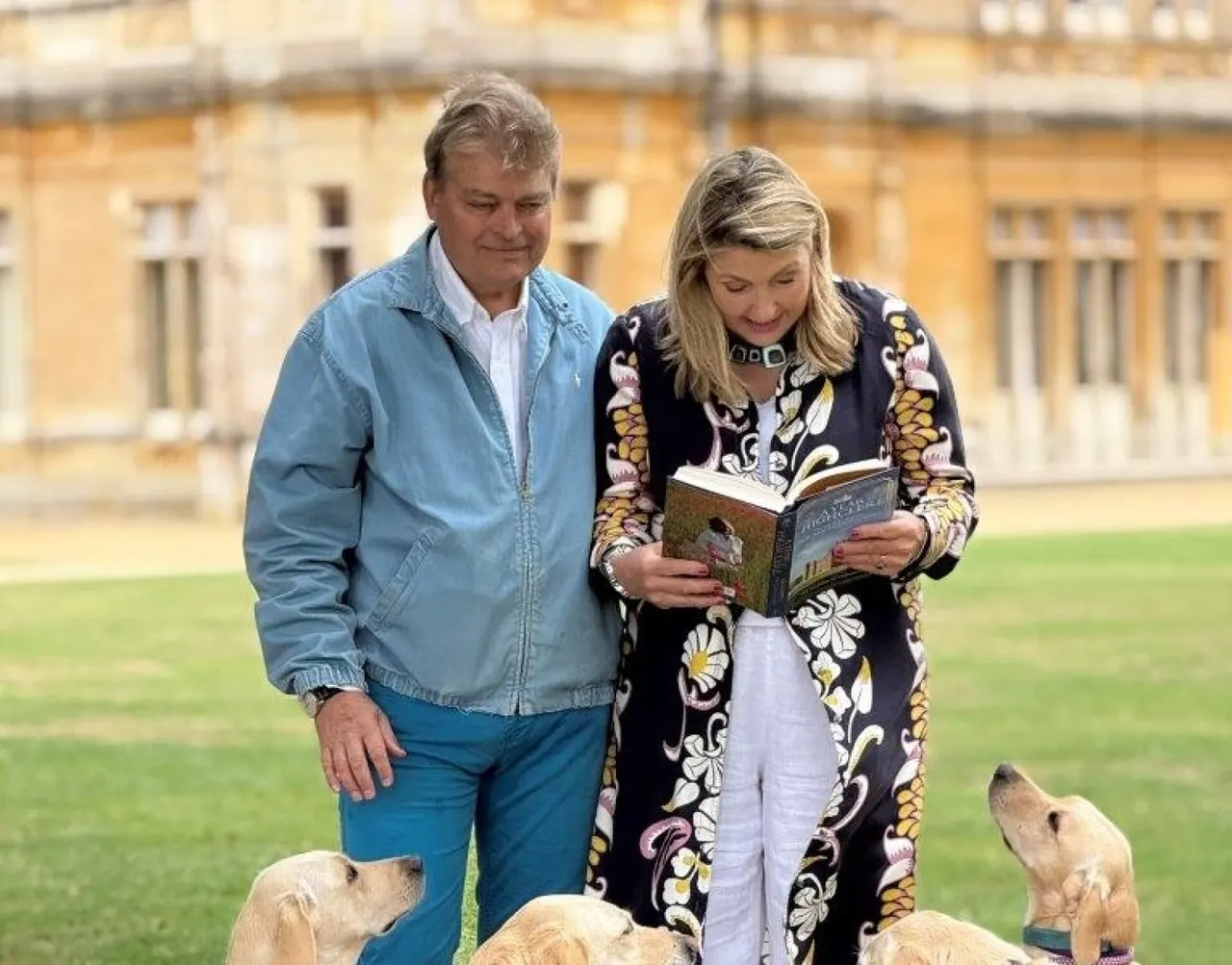A man and woman stand on a lawn in front of a stately stone building, the woman reading from a book whilst surrounded by dogs.