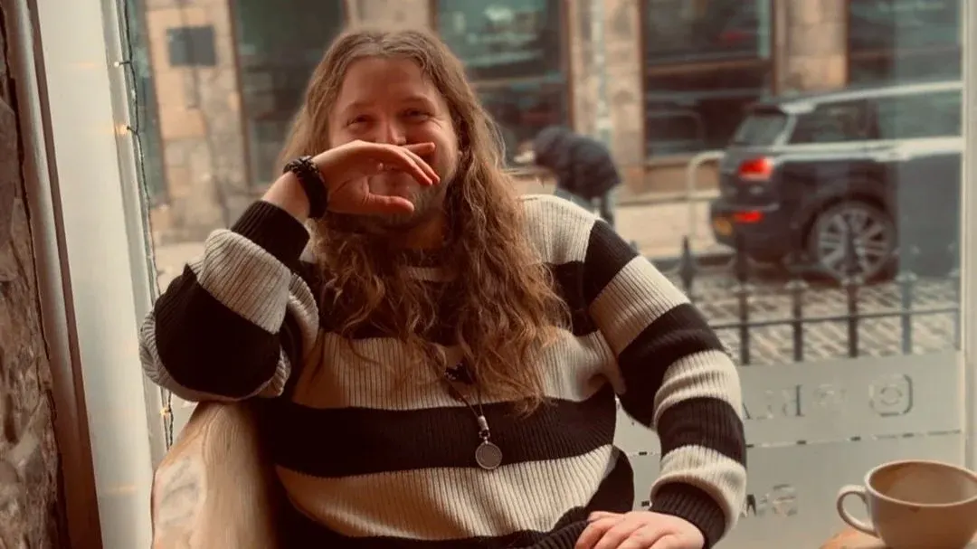 A smiling person with long hair sits by a window at a café, resting their chin on their hand with a cup of coffee on the table.