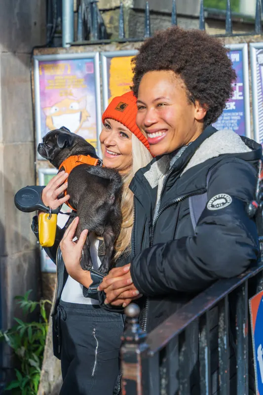Two people smiling whilst holding a small dog in winter clothing outdoors.