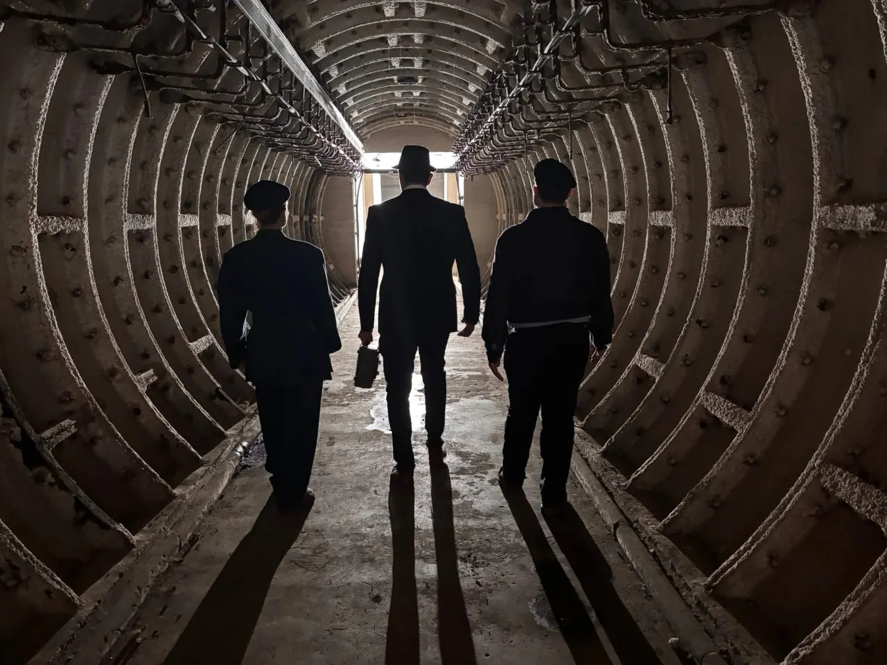 Three people in business attire walking through a long, cylindrical metal tunnel with arched ribbed walls.