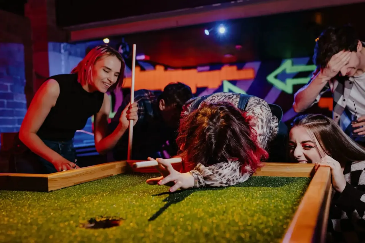 Young people playing mini golf under neon lighting in an indoor entertainment venue.